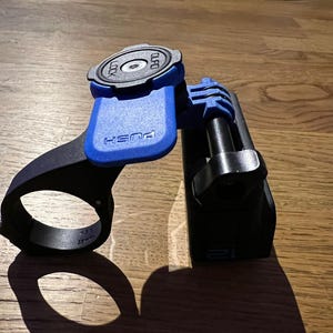 Op de afbeelding: Een zwarte en blauwe fietsstuurbevestiging met een druktoets om te ontgrendelen. De bevestiging heeft een ronde, zwarte basis met het woord "LOCK" erop.