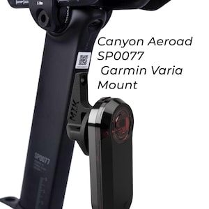 Puede incluir: Soporte negro Garmin Varia fijado a una tija de sillín de bicicleta negra. El soporte, con la etiqueta "MIK", sujeta una luz trasera Garmin Varia negra con lente roja. El texto en la tija del sillín dice "Canyon Aeroad SP0077".