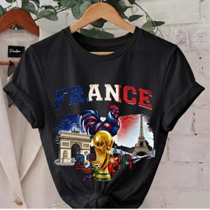 May include: Black t-shirt with a graphic featuring the word "FRANCE" in blue and red, a rooster, the Eiffel Tower, Arc de Triomphe, and a golden trophy. The design incorporates elements of the French flag.
