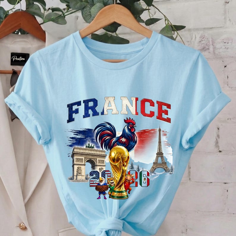 May include: Light blue t-shirt featuring the word "FRANCE" in red and white, with a rooster, the Eiffel Tower, and the Arc de Triomphe. A golden trophy and the numbers "2026" are also displayed. The shirt is on a wooden hanger.