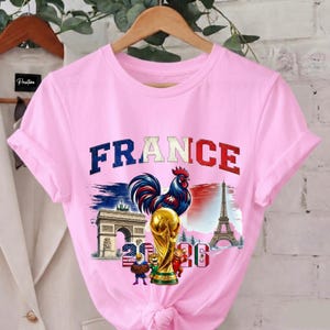 May include: Pink t-shirt with a graphic featuring the word "FRANCE" above a rooster, the Eiffel Tower, Arc de Triomphe, and a trophy. The graphic also includes the numbers "20" and "26".
