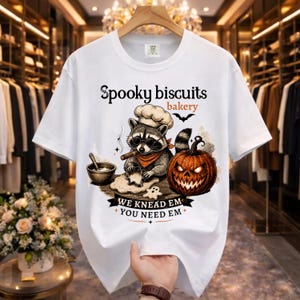 Peut inclure: Un t-shirt blanc avec une illustration de raton laveur portant une toque de chef et des lunettes, étalant de la pâte avec un emporte-pièce. Une citrouille sculptée est à côté. Texte : Spooky biscuits bakery, WE KNEAD EM YOU NEED EM.