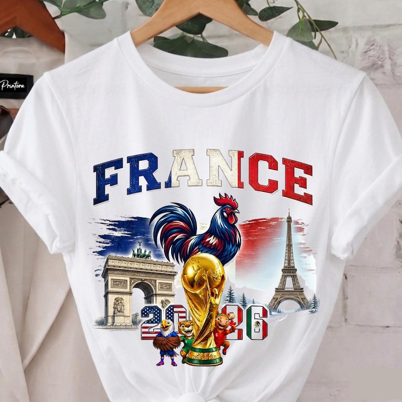 May include: White t-shirt featuring a graphic design with the word "FRANCE" in blue, white, and red. The design includes a rooster, the Eiffel Tower, the Arc de Triomphe, and a golden trophy. The shirt also has the numbers "20" and "26".