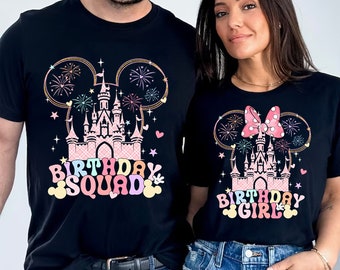 May include: Two black t-shirts featuring a pink castle graphic with fireworks and Mickey Mouse ears. The left shirt displays the text "BIRTHDAY SQUAD" in colorful letters, while the right shirt features a pink polka dot bow and "BIRTHDAY GIRL" text.