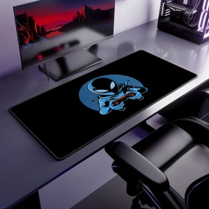 May include: Black rectangular gaming mouse pad featuring a blue circle with a cartoon astronaut holding a red game controller. The astronaut is wearing a blue suit. The mouse pad is on a desk with a computer monitor and gaming chair.
