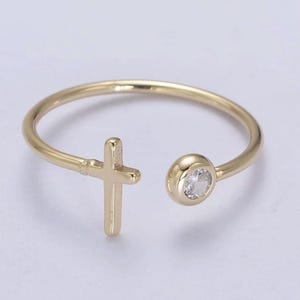 May include: A gold-colored ring with an open design. One end features a small cross, and the other end has a round setting with a clear, sparkling stone. The ring is likely made of gold-plated metal and is designed to be worn on a finger.