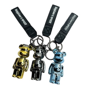 Op de afbeelding: Drie sleutelhangers met een metalen berenfiguur in goud, zilver en blauw. Elke sleutelhanger heeft een zwart leren bandje met de tekst "Attitude is everything" in wit.