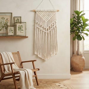 May include: A cream-coloured macrame wall hanging featuring intricate knotted patterns and long, flowing fringe suspended from a horizontal wooden dowel with a white cord loop for mounting, creating a textured geometric design on the wall.