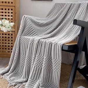 Puede incluir: Una manta de color gris claro con un patrón texturizado de rayas verticales acanaladas y cuadrados en relieve de punto de bodoque. La manta está colocada sobre una silla e incluye flecos decorativos a lo largo del borde inferior.