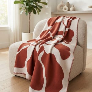 May include: A knit throw blanket featuring a large-scale abstract floral pattern in rust orange and cream. The blanket has a whip-stitched edge and a small brown leather-like patch with a W logo. The text TEXTURED COMFORT is visible above.