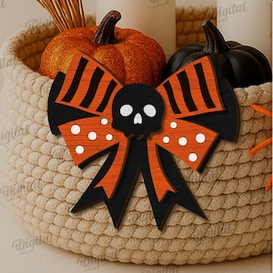 K&ouml;nnte beinhalten: Eine dekorative Halloween-Schleife mit einem Totenkopf in der Mitte, orangefarbenen und schwarzen Streifen und wei&szlig;en Punkten. Die Schleife befindet sich vor einem geflochtenen Korb mit K&uuml;rbissen.