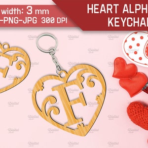 May include: A wooden heart-shaped keychain with a decorative letter F cut out. The keychain is made of light brown wood and has a silver key ring attached. The background is pink.