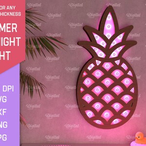 Puede incluir: Una luz nocturna de piña de madera rosa con luces LED. La piña está en una pared rosa con una hoja de palma en el fondo.