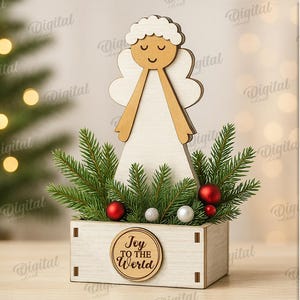 May include: A wooden Christmas angel decoration with a light wood body and wings, and a smiling face. The angel is set in a small wooden box with greenery, red and silver ornaments, and the text "Joy to the World".