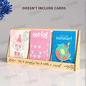 May include: A wooden card display stand holding three greeting cards with colorful illustrations. The cards feature a cartoon fox, pink birthday decorations, and a frog with a popsicle. The stand has the text "Say it simply. Say it with a card."