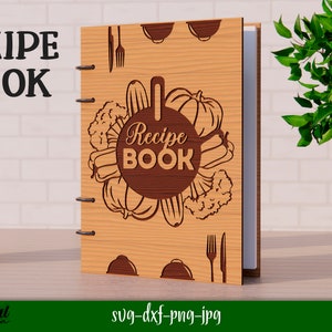 Może przedstawiać: Drewniana okładka książki kucharskiej z brązowym tłem i k&oacute;łkiem z tekstem "Recipe Book" otoczonym warzywami. Okładka ma r&oacute;wnież widelec i n&oacute;ż w prawym dolnym rogu.