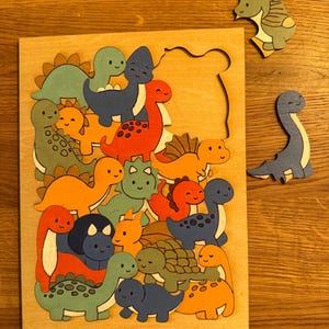 Puede incluir: Un rompecabezas de madera con una colecci&oacute;n de dinosaurios coloridos estilo caricatura en tonos naranja, azul y verde. El tablero tiene un &aacute;rea recortada rectangular, con dos piezas individuales con forma de dinosaurio a la derecha.