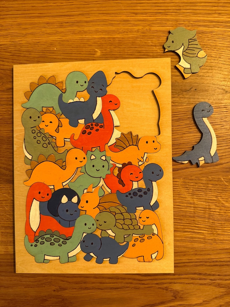 Puede incluir: Un rompecabezas de madera con una colecci&oacute;n de dinosaurios coloridos estilo caricatura en tonos naranja, azul y verde. El tablero tiene un &aacute;rea recortada rectangular, con dos piezas individuales con forma de dinosaurio a la derecha.