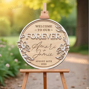 May include: A wooden wedding sign on an easel. The sign is round with the words "WELCOME - TO OUR - FOREVER" and the names "Amari & Jamie" with the date "AUGUST 20TH 2026". The sign is decorated with floral designs.
