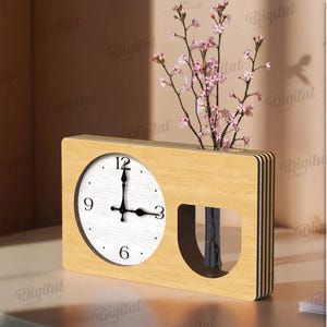 K&ouml;nnte beinhalten: Eine Kombination aus Holz-Uhr und Vase. Das Zifferblatt ist wei&szlig; mit schwarzen Zeigern und Ziffern. Der Vasenbereich enth&auml;lt rosa bl&uuml;hende Zweige. Die Uhr steht auf einem wei&szlig;en Tisch.