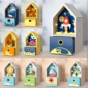 Op de afbeelding: Een verzameling van tien houten huisvormige dozen met lades, elk met een uniek ontwerp. De dozen zijn in verschillende kleuren geschilderd, met thema's als een walvis, bijen, een raket en een regenboog. De materiaalbreedte is 3 mm.