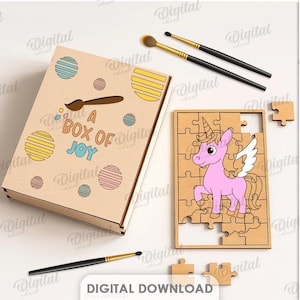 May include: A wooden jigsaw puzzle with a pink unicorn design, and a box with the text "A BOX OF JOY". The box is decorated with colorful circles and a paintbrush. Two paintbrushes are also visible.