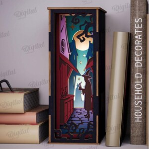 May include: A laser-cut wooden Halloween-themed shadow box depicting a witch in a spooky alleyway. The box is black with a 3 mm width and features a witch, buildings, and a full moon. A black spider sits on a stack of books. The text "HOUSEHOLD DECORATES" is visible.