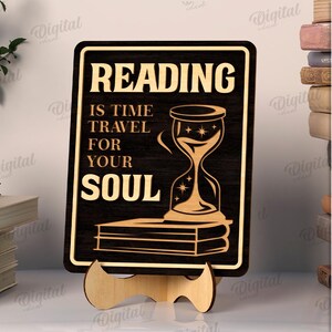 K&ouml;nnte beinhalten: Schwarzes Holzschild mit dem Text "READING IS TIME TRAVEL FOR YOUR SOUL" und einer Sanduhr-Illustration. Das Schild wird auf einem Holzst&auml;nder pr&auml;sentiert. Die Breite des Schildes betr&auml;gt 3 mm. Ein Stapel B&uuml;cher und ein Granatapfel sind im Hintergrund.