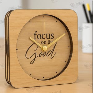 K&ouml;nnte beinhalten: Eine quadratische Holzuhr mit hellbraunem Finish. Das Zifferblatt zeigt die Worte "focus on the Good" in einer Schriftart. Die Uhrzeiger sind goldfarben.