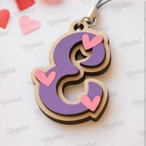 May include: A layered wooden charm in the shape of the letter "E", painted purple with pink heart accents. The charm is attached to a black cord. The background includes pink and red heart shapes and a pink ribbon.