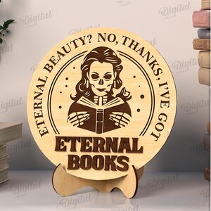 K&ouml;nnte beinhalten: Ein rundes Holzschild mit dem Text "Eternal Beauty? No, Thanks, I've Got Eternal Books." Das Schild zeigt eine Grafik einer sch&auml;delgesichtigen Frau, die ein Buch liest. Das Schild wird auf einem Holzst&auml;nder ausgestellt.
