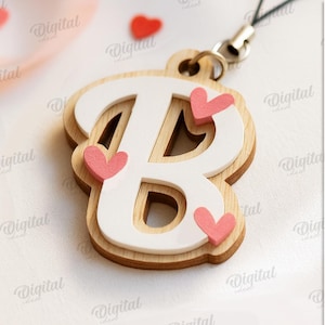 May include: A wooden letter "B" charm with a white overlay and pink heart accents. The charm is attached to a black cord. The image also includes the text "SUITED FOR ANY MATERIAL THICKNESS" and file type options.