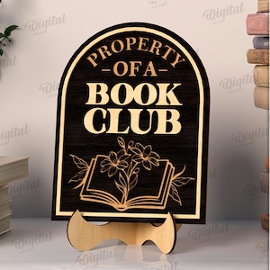 K&ouml;nnte beinhalten: Ein 3 mm dickes Holzschild mit dem Text "PROPERTY OF A BOOK CLUB" in einem dekorativen Bogen. Das Schild zeigt ein offenes Buch mit floralen Akzenten. Es wird von einem Holzst&auml;nder getragen.