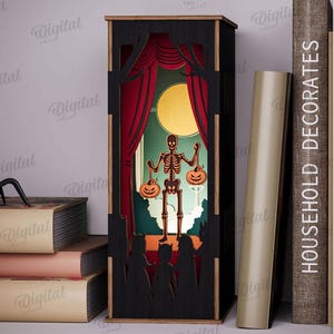 May include: A black Halloween-themed shadow box with a skeleton holding jack-o'-lanterns on a stage with red curtains and a full moon. A black spider sits on a stack of books. The text "HOUSEHOLD DECORATES" is on a book.