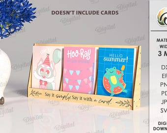 May include: A wooden card display stand holding three greeting cards with colorful illustrations. The cards feature a cartoon fox, pink birthday decorations, and a frog with a popsicle. The stand has the text "Say it simply. Say it with a card."