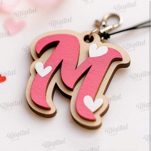 May include: A wooden keychain featuring the letter "M" in pink, adorned with white heart accents. The keychain has a natural wood border and a silver-colored clasp. The background includes scattered heart-shaped confetti and a ribbon.