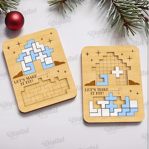 May include: Two wooden puzzle game pieces with a Tetris-like design, featuring blue and white blocks. The phrase "LET'S MAKE IT FIT!" is printed on each piece. A red ornament and evergreen branches are in the background. Digital download.