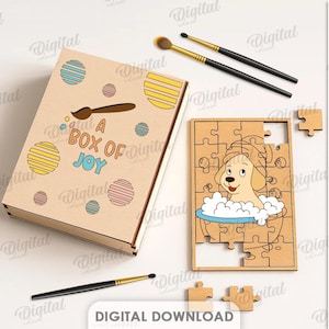 May include: A wooden puzzle and box set with the text "A Box of Joy". The box is decorated with colourful circles and a paintbrush. The puzzle features a cartoon dog in a bathtub. Includes paintbrushes.