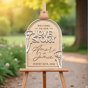 May include: Wooden wedding sign on an easel, featuring the words "Welcome to our Love Story" and the names "Amari & Jamie" with the date August 20th, 2026. The sign has a decorative floral design and is set outdoors.