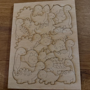 Puede incluir: Una tabla rectangular de madera clara con contornos de dinosaurios de dibujos animados grabados con l&aacute;ser. El dise&ntilde;o incluye varias especies con caras sonrientes, manchas y caparazones texturizados, dispuestos en un patr&oacute;n denso y superpuesto.