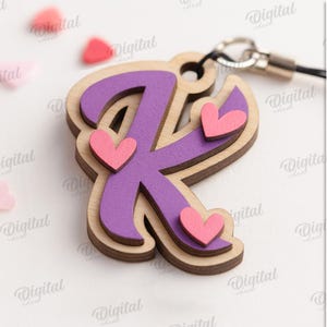 May include: A layered wooden letter "K" charm with a purple center and pink hearts. The charm is attached to a silver-colored clasp. The image also includes scattered heart-shaped confetti and the text "SUITED FOR ANY MATERIAL THICKNESS".