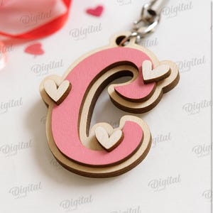 May include: A wooden letter "C" keychain with a pink center and light wood border, adorned with small heart accents. The keychain is on a silver chain, with the text "SUITED FOR ANY MATERIAL THICKNESS" and file type options on the right.