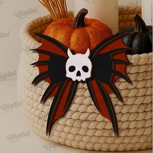 K&ouml;nnte beinhalten: Eine Halloween-Dekorationsschleife mit einem Totenkopf in der Mitte und einem Fledermausfl&uuml;gel-Design in Schwarz, Rot und Holzt&ouml;nen. Die Schleife befindet sich in einem geflochtenen Korb mit K&uuml;rbissen und Kerzen. Der Text lautet "SUITED FOR ANY MATERIAL THICKNESS".