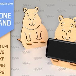 May include: A phone stand made of wood, shaped like a cartoon animal with a smiling face. The stand is designed to hold a smartphone upright. The text "PHONE STAND" is visible in the image.