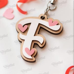 May include: Wooden letter "F" keychain with a white center and pink heart accents. The keychain is layered with a light brown wood and includes a silver-colored metal ring. The image also includes pink heart shapes and red ribbon.