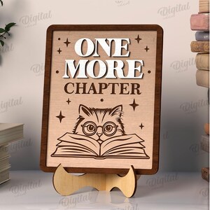 K&ouml;nnte beinhalten: Holzschild mit den Worten "ONE MORE CHAPTER" und einer Katze mit Brille, die ein Buch liest. Das Schild steht auf einem Holzst&auml;nder. Das Schild ist 3 mm breit. Digitaler Download.