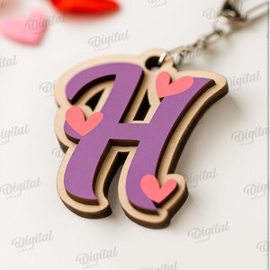 May include: A wooden keychain featuring the letter "H" in purple with pink hearts. The keychain has a layered design with a light brown border. The design is attached to a silver chain. The image also includes the text "SUITED FOR ANY MATERIAL THICKNESS".