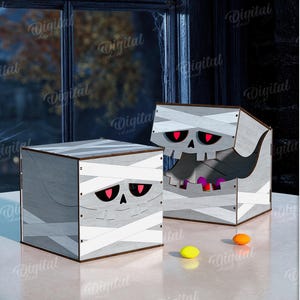 K&ouml;nnte beinhalten: Zwei h&ouml;lzerne Halloween-Boxen, jede mit einem Mumien-Design. Die Boxen sind grau mit wei&szlig;en Bandagedetails und roten Augen. Die Boxen stehen auf einer wei&szlig;en Oberfl&auml;che mit einem K&uuml;rbis und S&uuml;&szlig;igkeiten im Hintergrund.