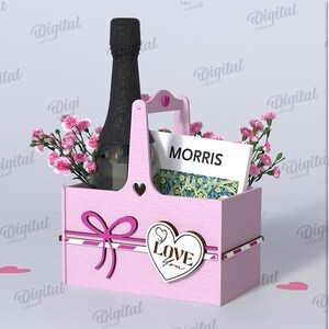 Könnte beinhalten: Ein rosa Holzgeschenkkorb mit Griff, der eine Champagnerflasche, Blumen und eine Karte mit der Aufschrift "MORRIS" enthält. Der Korb hat eine Schleife und ein Herz mit "Love You". Die Materialbreite beträgt 3 mm.