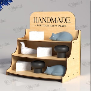 May include: A three-tiered wooden display shelf with the text "HANDMADE - FOR YOUR HAPPY PLACE." The shelf holds decorative items, including white cubes, gray cat figurines, and black mushroom-shaped objects. The material width is 3 mm.